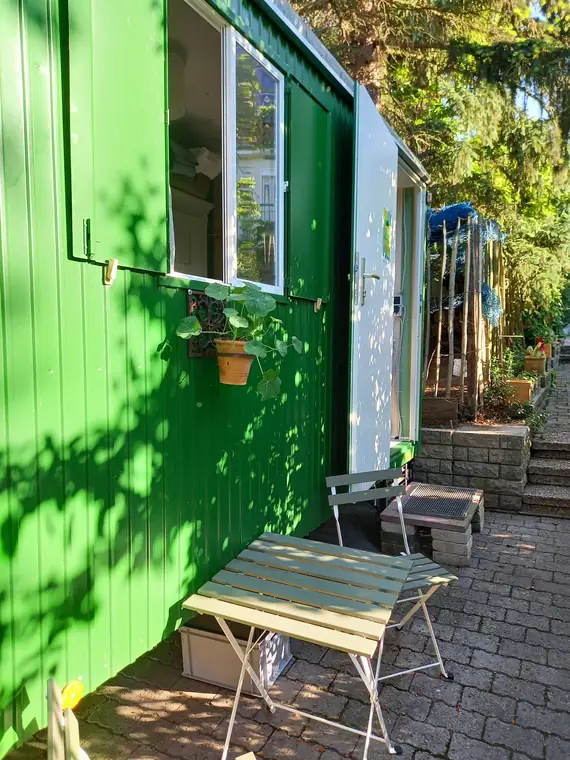 Grüne Bauwagenwand mit Fenster und Blumentopf, davor eine Holzbank