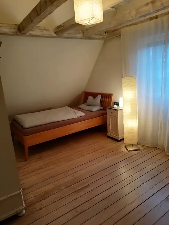 Holzbett unter der Dachschräge, daneben eine Stehleuchte und ein Fenster mit Vorhang