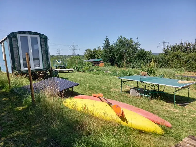 Bauwagen mit großer Fensterfront auf der Wiese, davor Solarpanel, Tischtennisplatte und zwei Kajaks