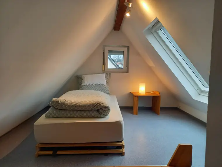 Bett unter der Dachschräge neben dem Dachfenster, daneben eine Leuchte